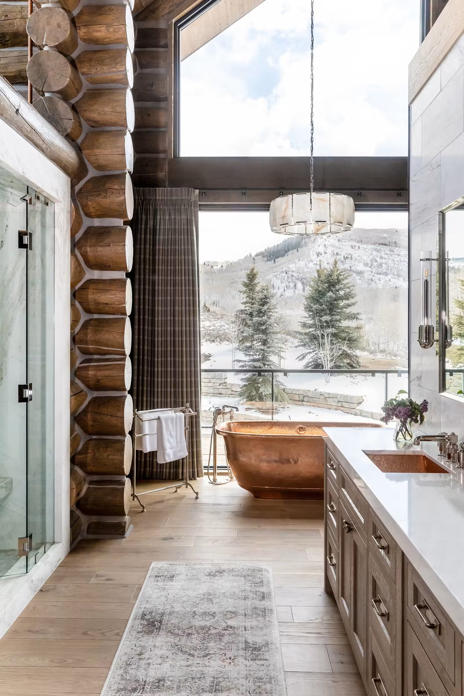 Log home bathroom with light wood flooring, freestanding copper bathtub, and mountain views.