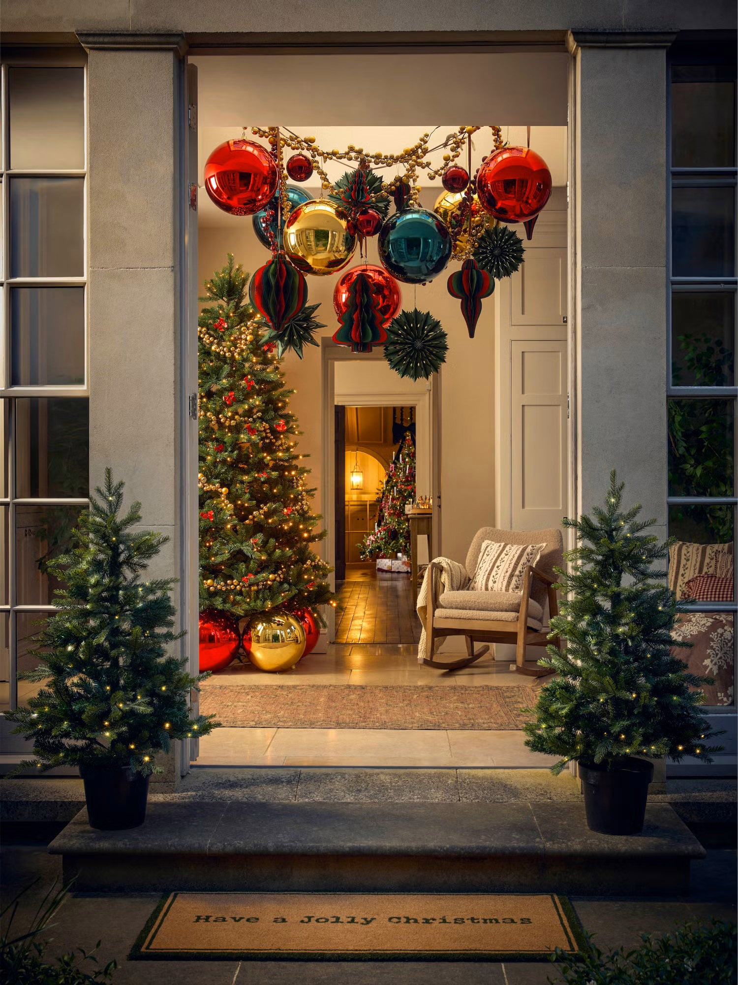 Front porch with a Christmas doormat and two pre-lit trees in black pots, with a decorated tree visible through the glass door