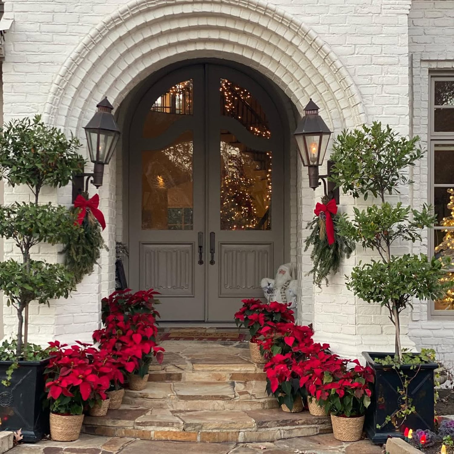 Red poinsettias in wicker baskets lining white brick porch steps beside topiaries and lanterns with red bows