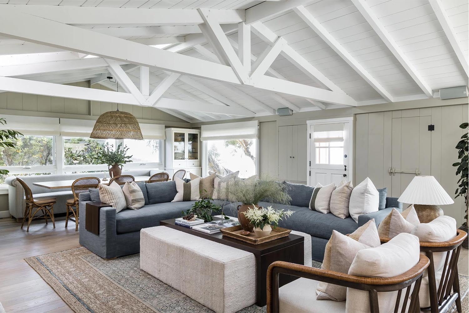 Bright coastal living room with light oak flooring, vaulted white ceiling, gray-blue sectional, woven chairs, and textured area rug showing a layered, neutral color palette.