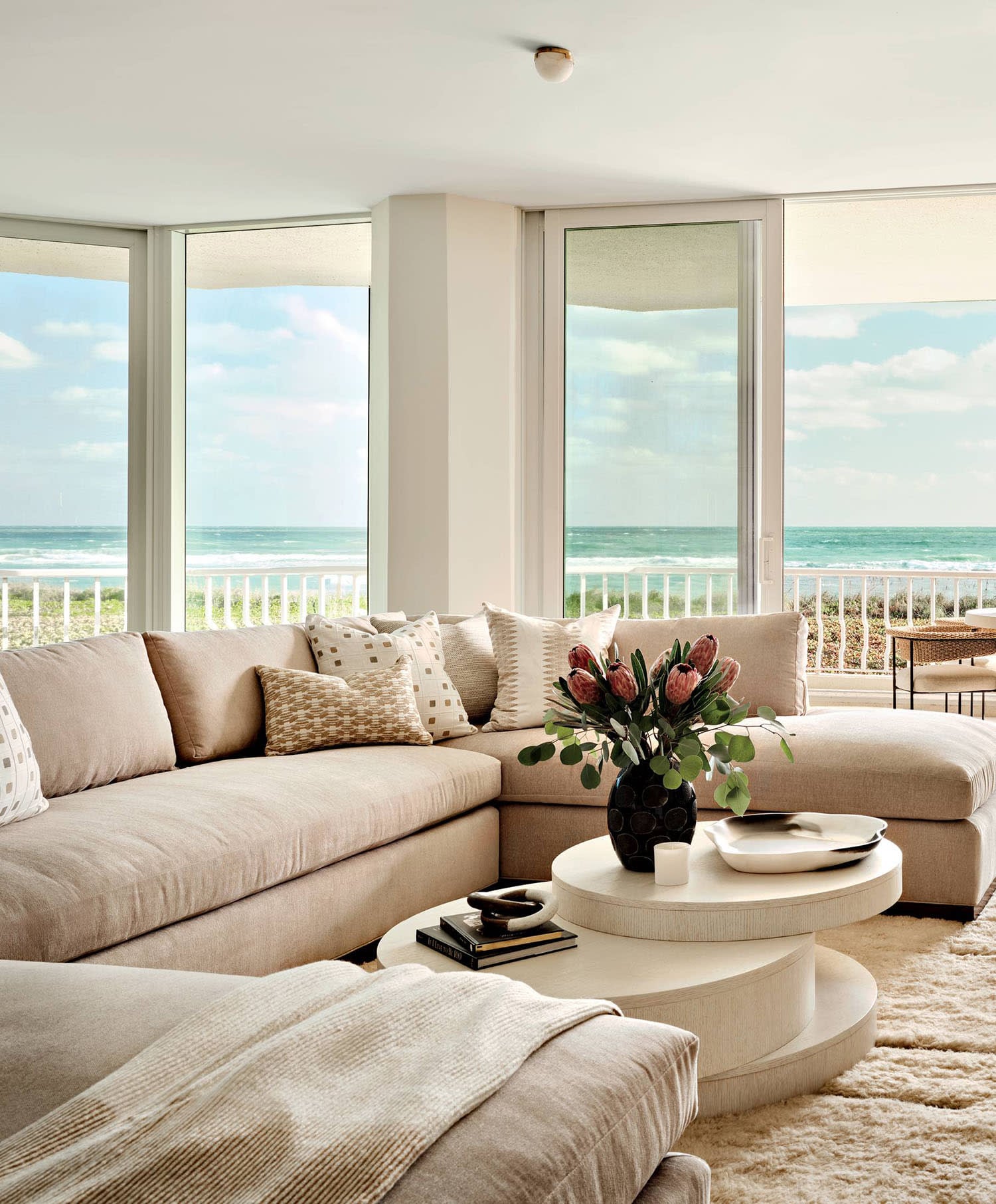 Modern coastal living room in neutral sandy tones with floor-to-ceiling windows framing ocean and sky views, large sectional, whitewashed circular coffee table, and layered pillows and throws.